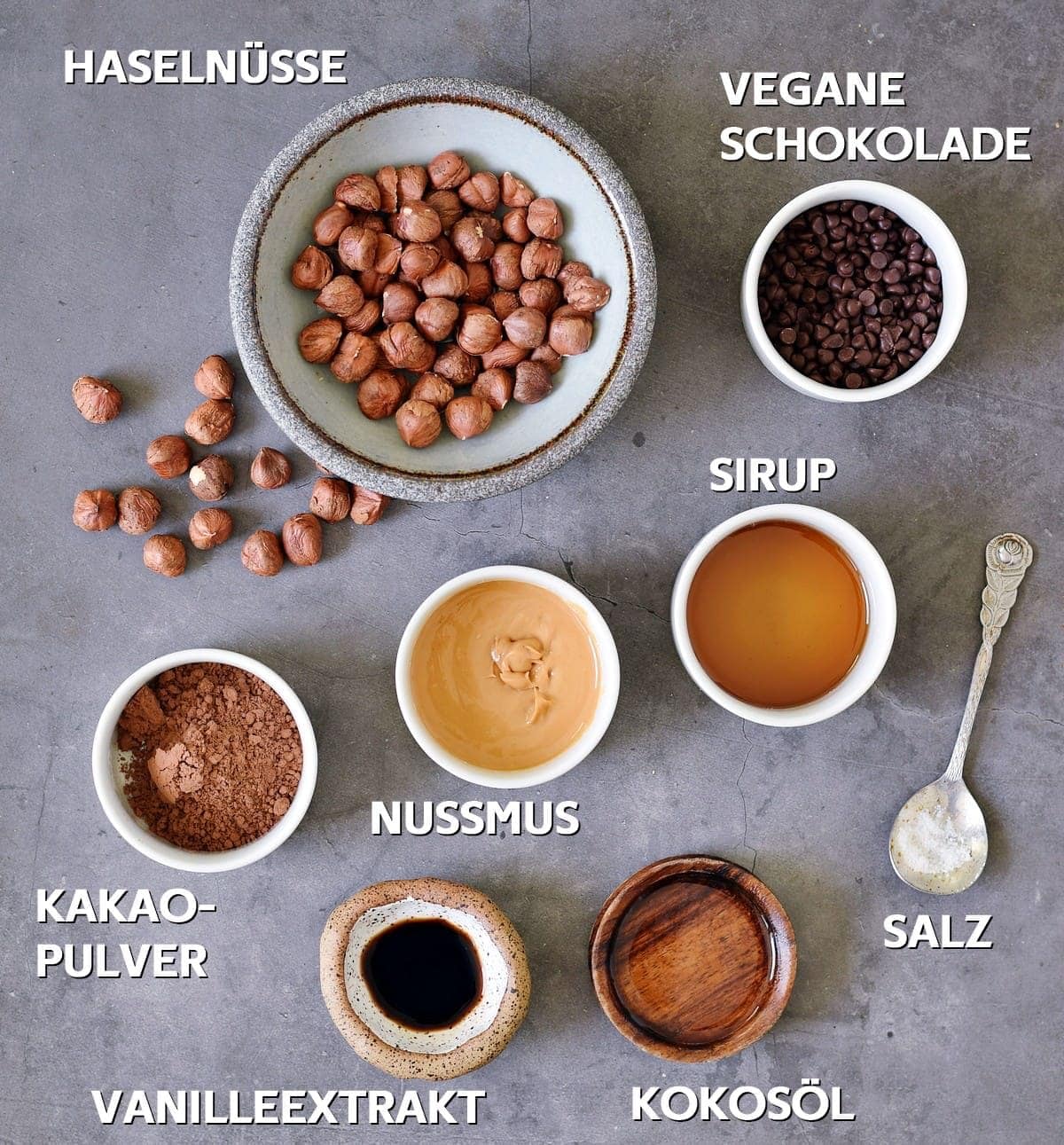 Vegane Schokolade, Sirup, Salz, Kokosöl, Nussmus, Vanilleextrakt, Kakaopulver, Haselnüsse auf grauem Hintergrund