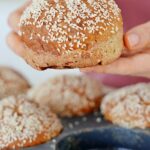 Hände halten glutenfreies Burgerbrötchen über grauer Backform mit den selben Brötchen