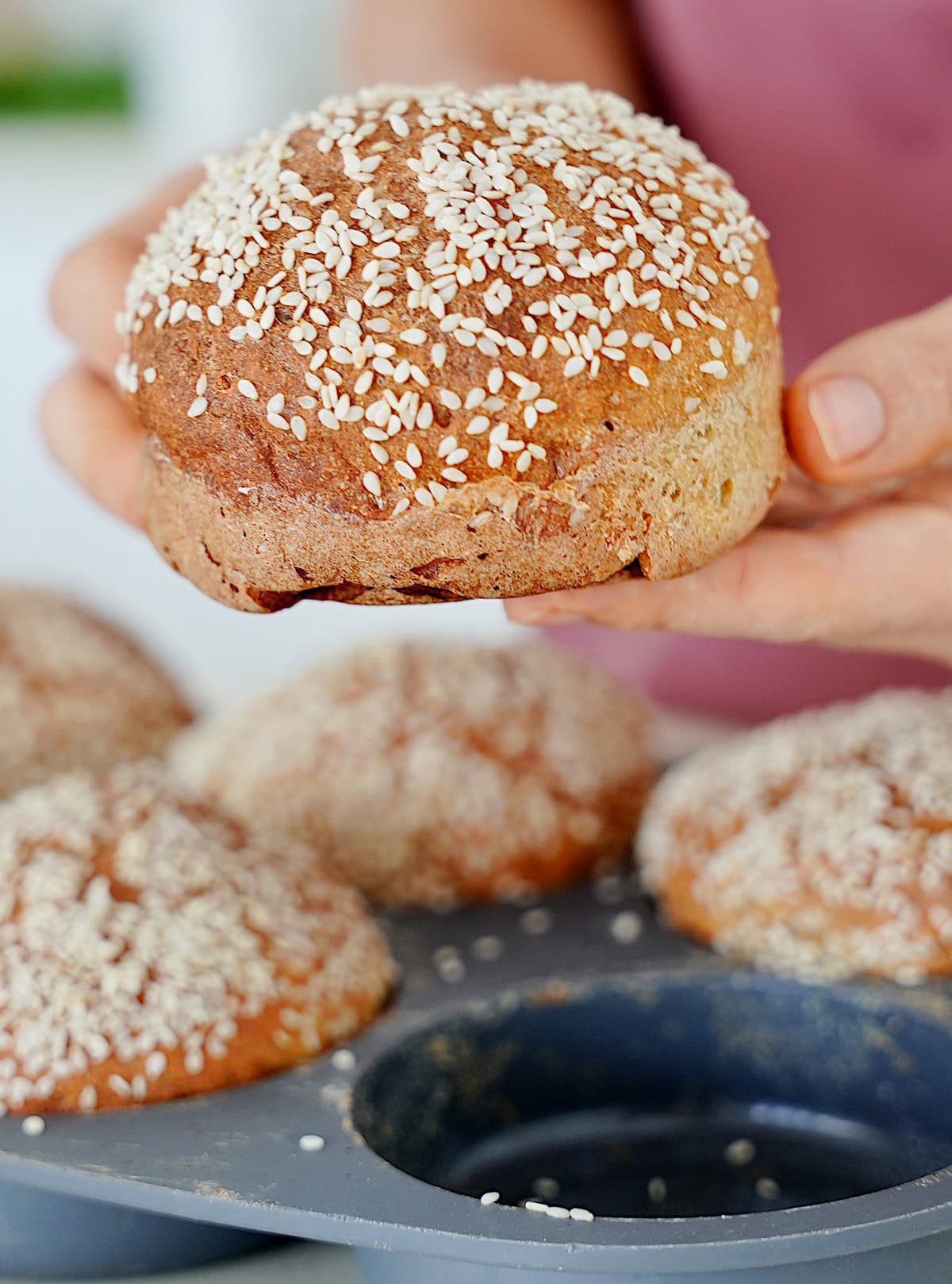 Hände halten glutenfreies Burgerbrötchen über grauer Backform mit den selben Brötchen