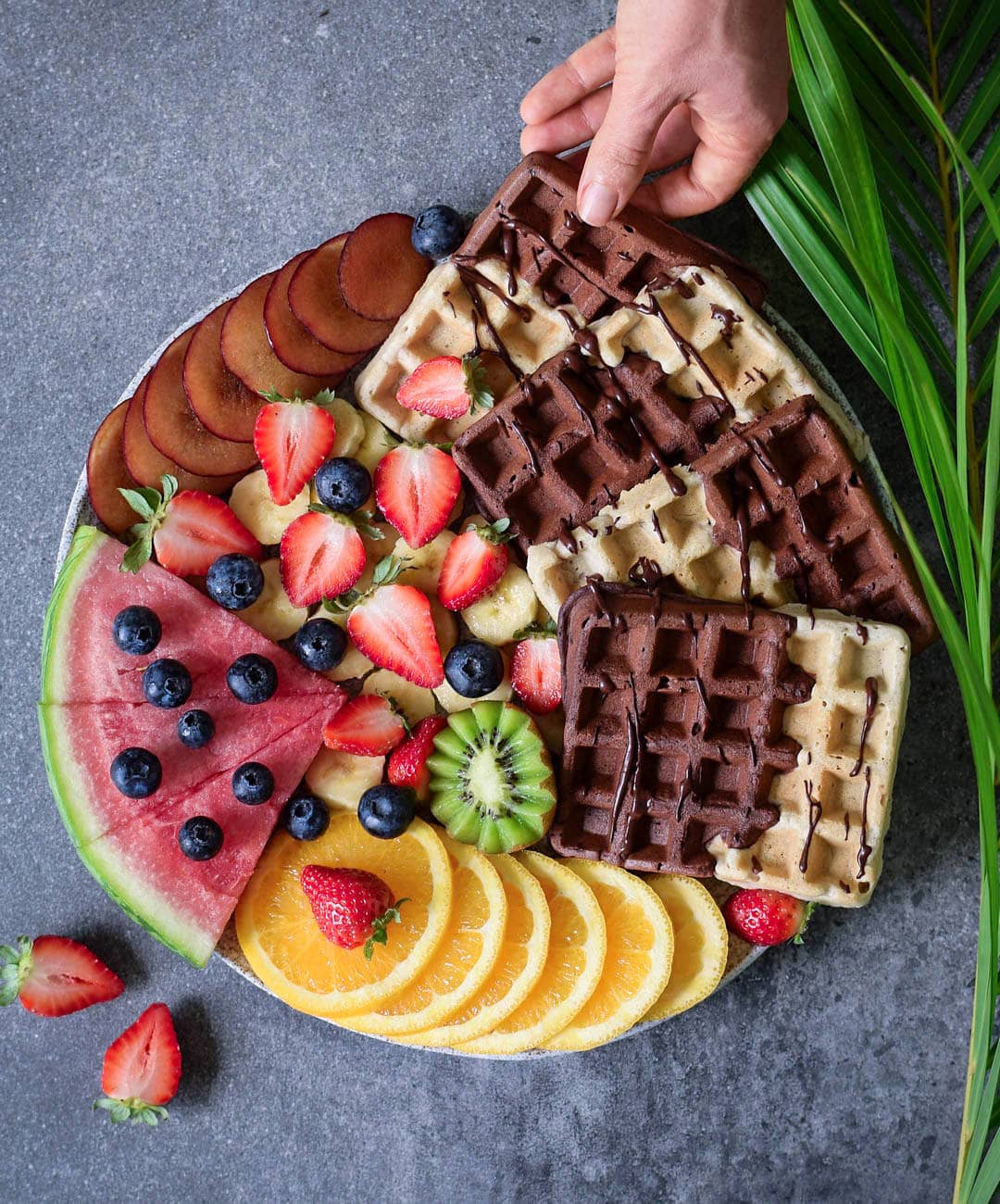 Hand grabbing homemade gluten-free vegan waffles with fruits, a chocolate sauce, arranged on a board