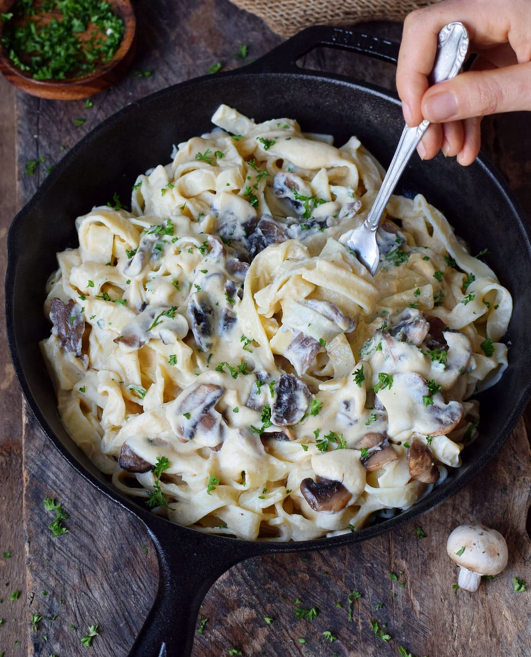 Hand holding fork submerged in vegan Alfredo Fettuccine with mushrooms in a skillet