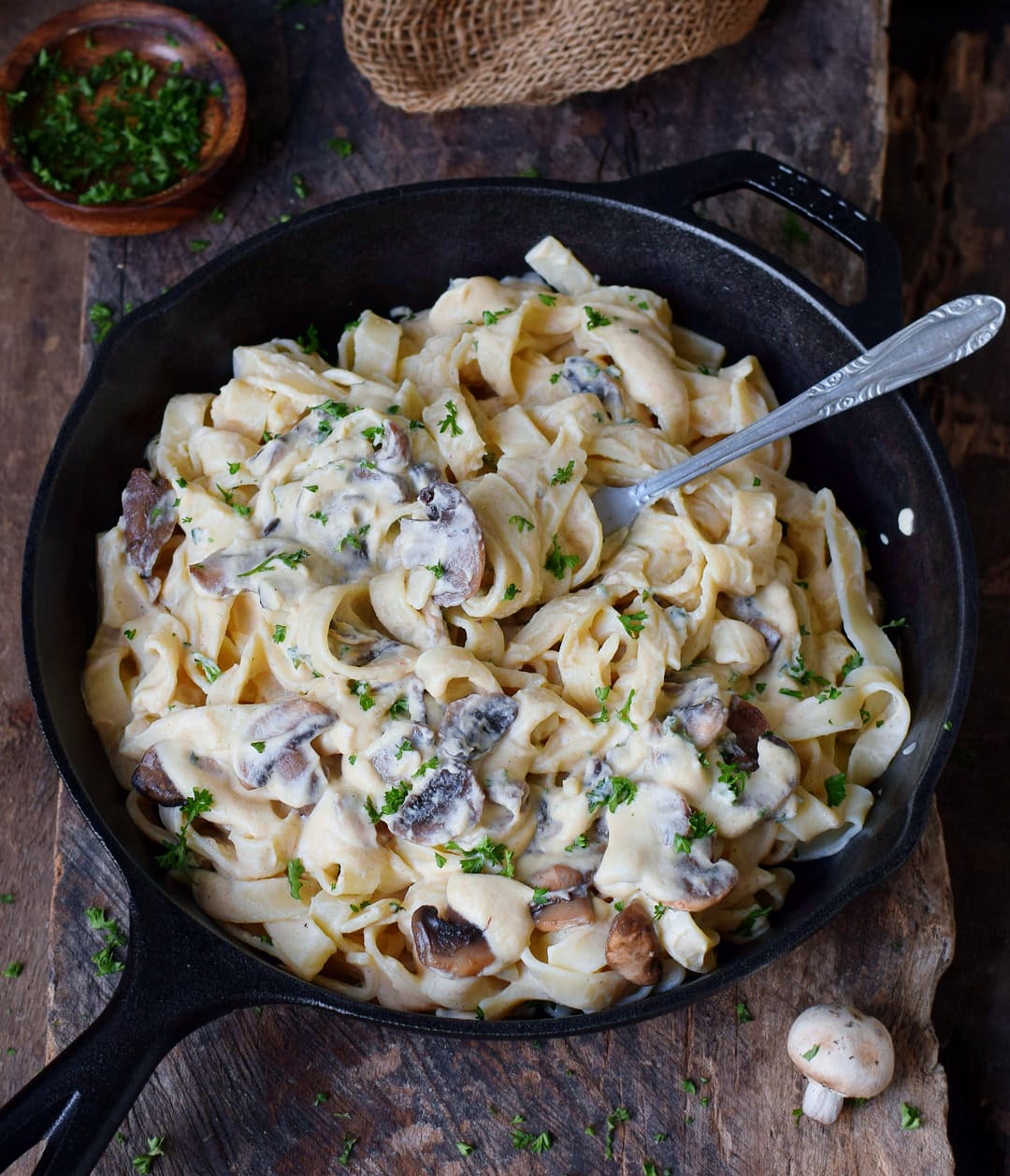 Fork submerged in vegan Alfredo sauce with mushrooms in a skillet