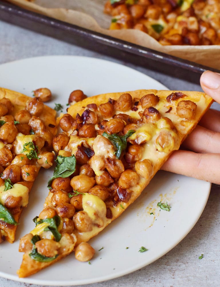 Hand hält ein Stück Pizza aus Süßkartoffeln belegt mit Kichererbsen und veganem Käse