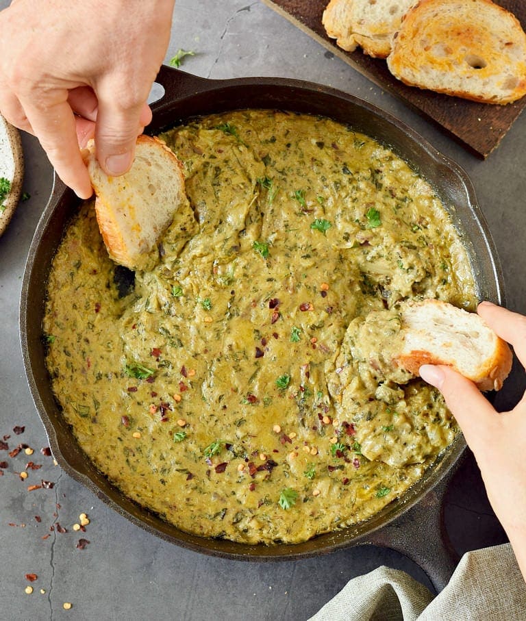 two hands submerge baguette into a spinach artichoke dip