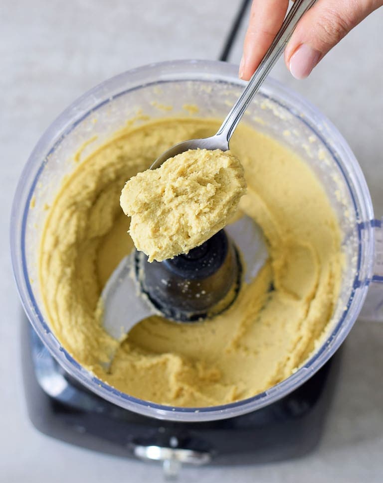 hand holding a spoon with creamy chickpea cheese above a food processor