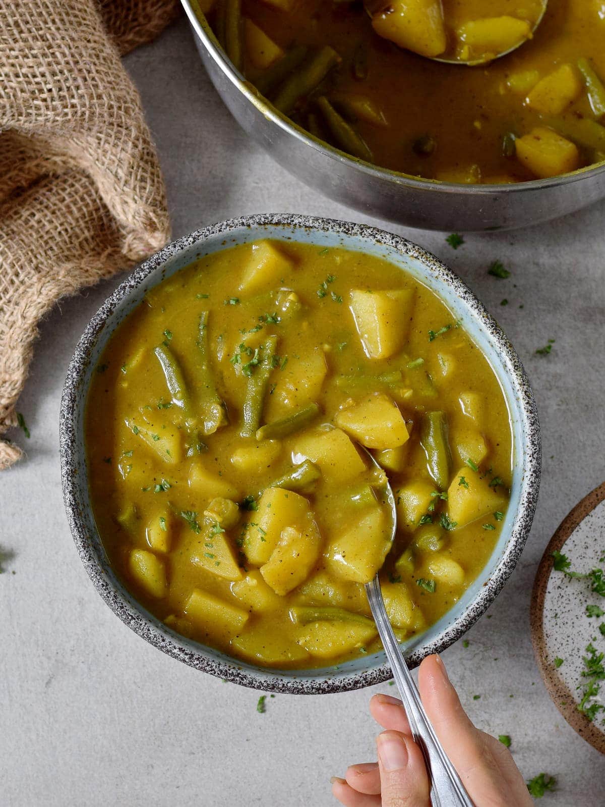 Veganes Kartoffel-Gemüse Curry mit grünen Bohnen in einer Schale und Löffel in einer Hand