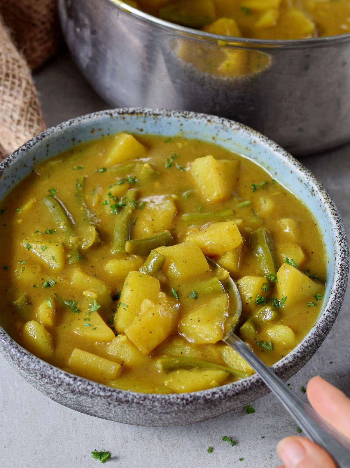 Veganes Kartoffel-Curry mit grünen Bohnen in einer Schale und Löffel in einer Hand