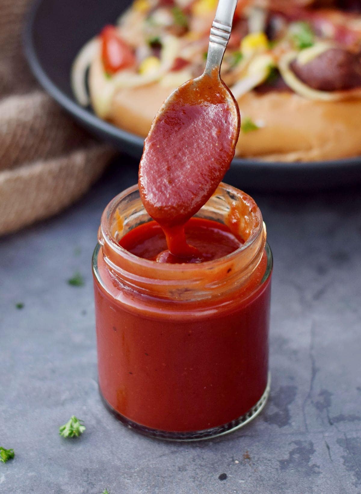 BBQ Sauce im Glas mit Löffel und veganen Hot Dogs im Hintergrund