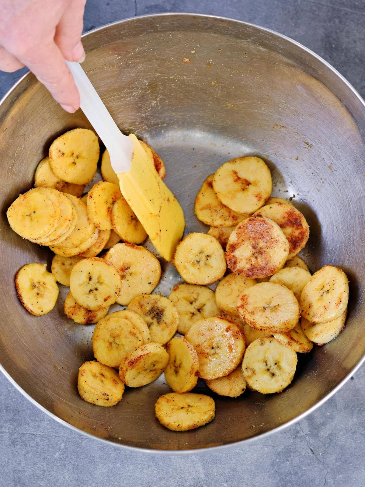 stiring plantain slices with a rubber spatula in silver bowl