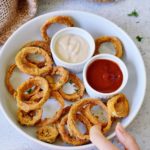 hand grabbing vegan onion rings in a bowl with 2 dips