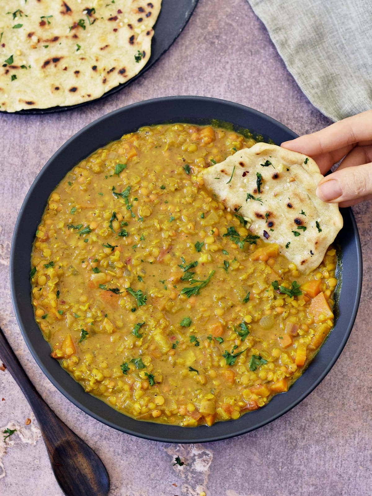 Hand dipping a piece of vegan naan bread into red lentil dahl in black bowl