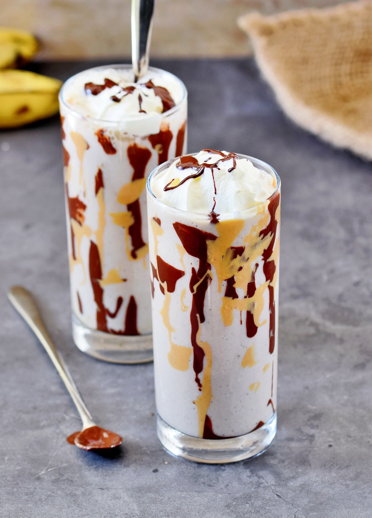 banana shake decorated with vegan whipped cream, chocolate and peanut butter in 2 glasses with long spoons