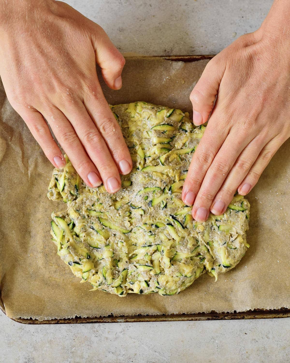Hände verteilen Pizzateig aus Zucchinis auf geöltem Backpapier.