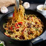 garlic noodles in black skillet with veggies and tongs