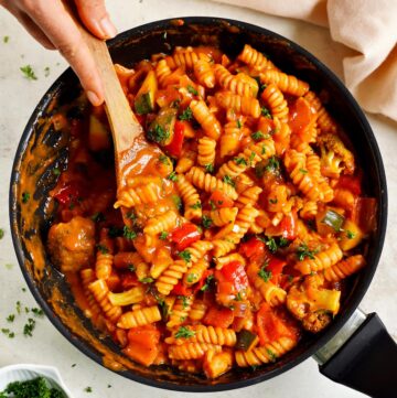 hand stirring creamy tomato sauce vegetable pasta in black skillet