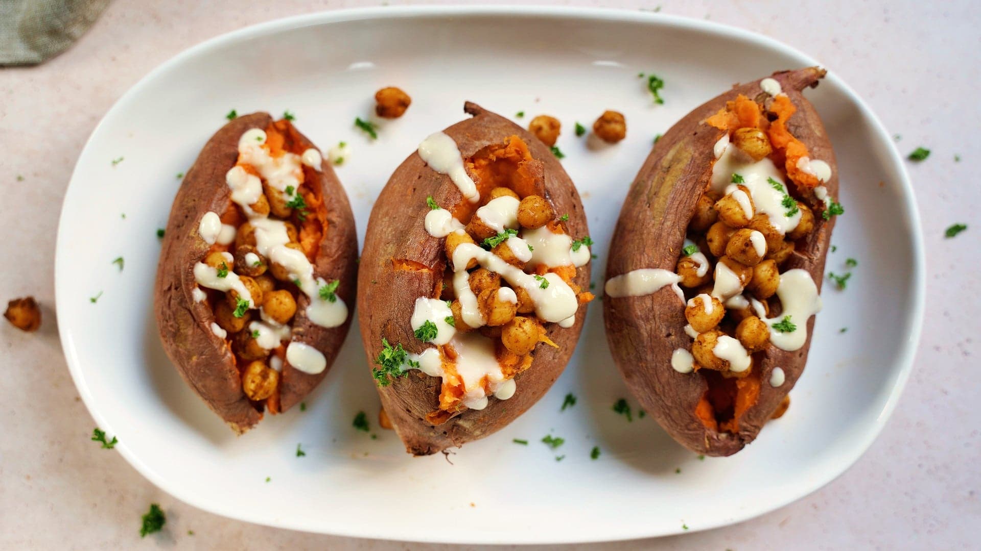 horizontal shot of 3 baked sweet potatoes on a white plate with roasted chickpeas and white sauce