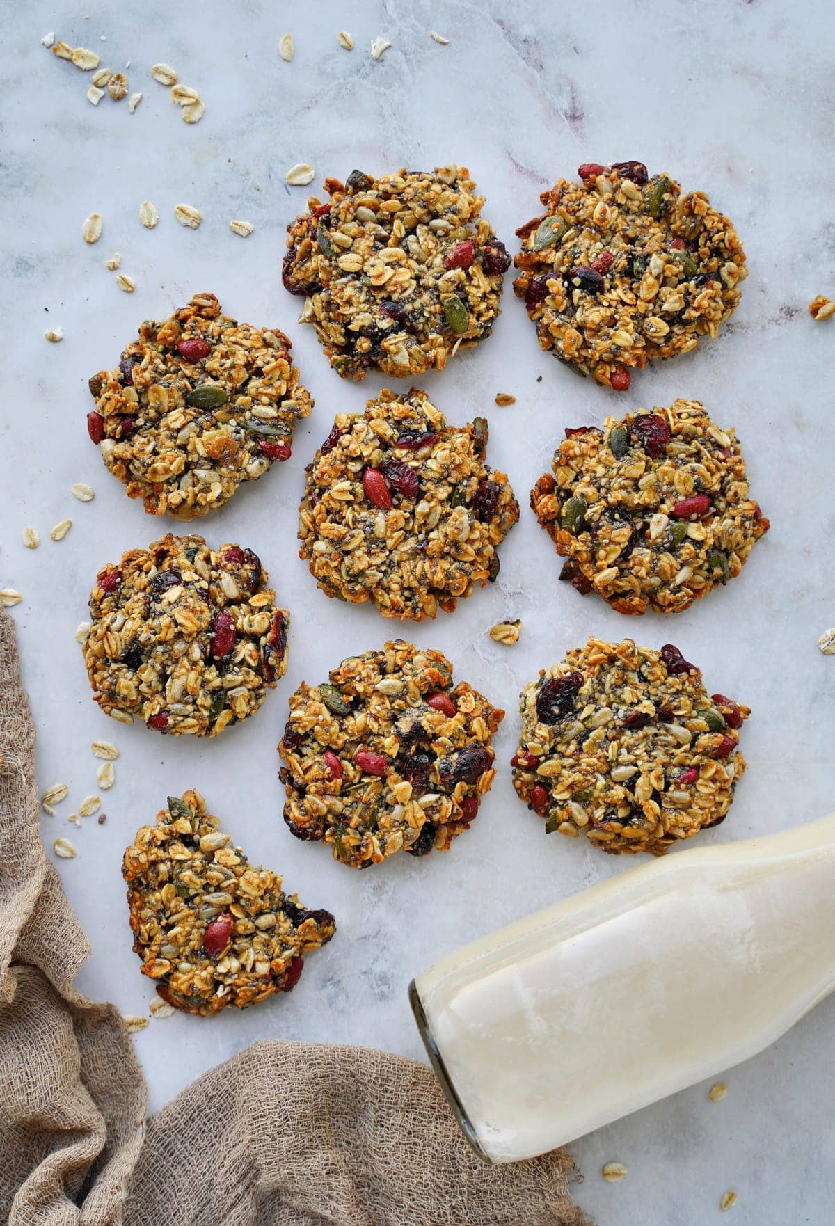 9 healthy cookies with oats, seeds, and dried fruit on white marble with milk bottle