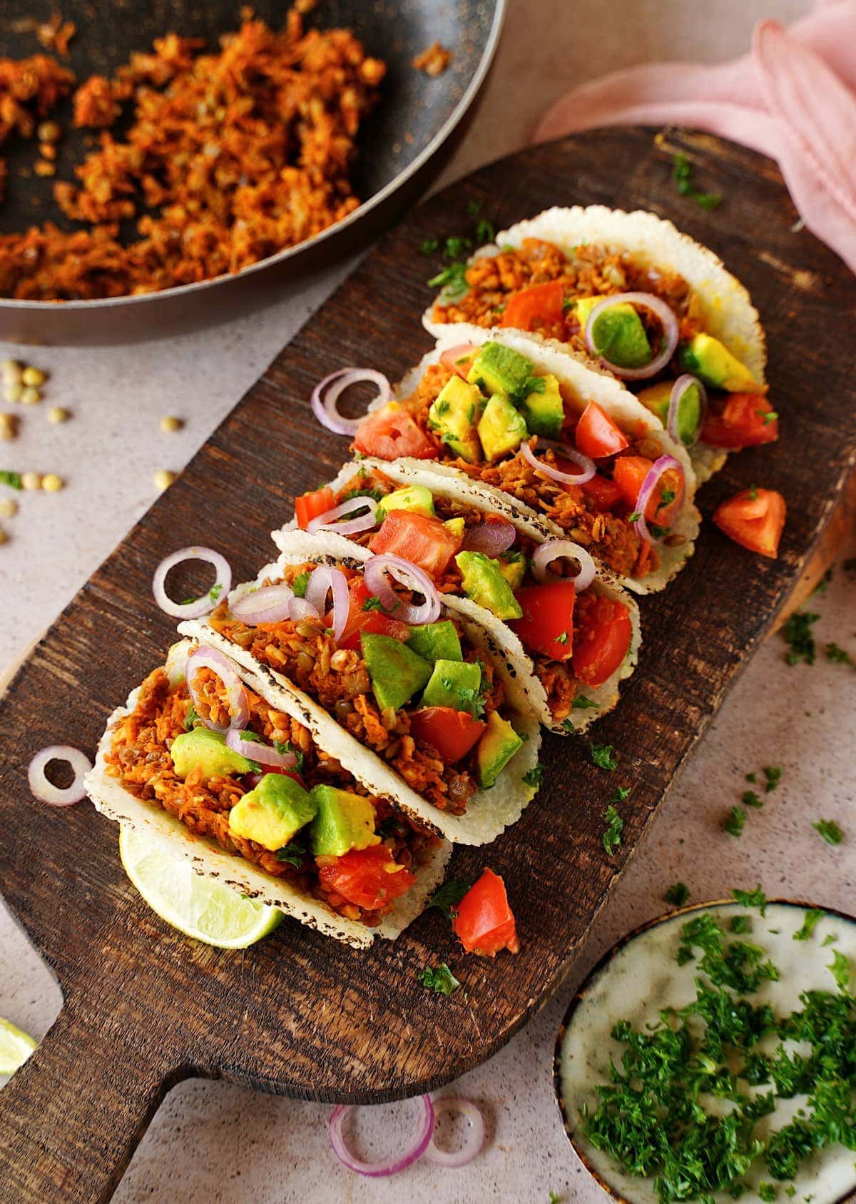 tortillas with lentil taco meat topped with tomato, onion, and avocado on wooden board