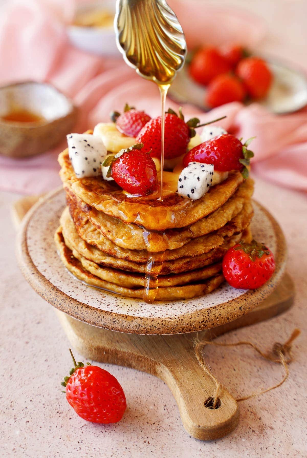 spoon drizzling maple syrup over a stack of protein pancakes with fruits