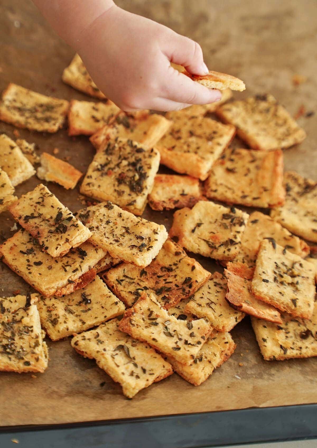 A childs hand reaches for a square cracker covered in herbs on a baking sheet
