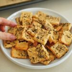 A hand reaches for a square herb-seasoned chickpea cracker on a white plate full of similar crackers