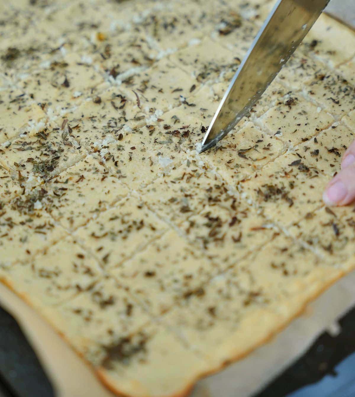 A hand slices a large, seasoned chickpea flatbread dough with herbs into squares using a knife