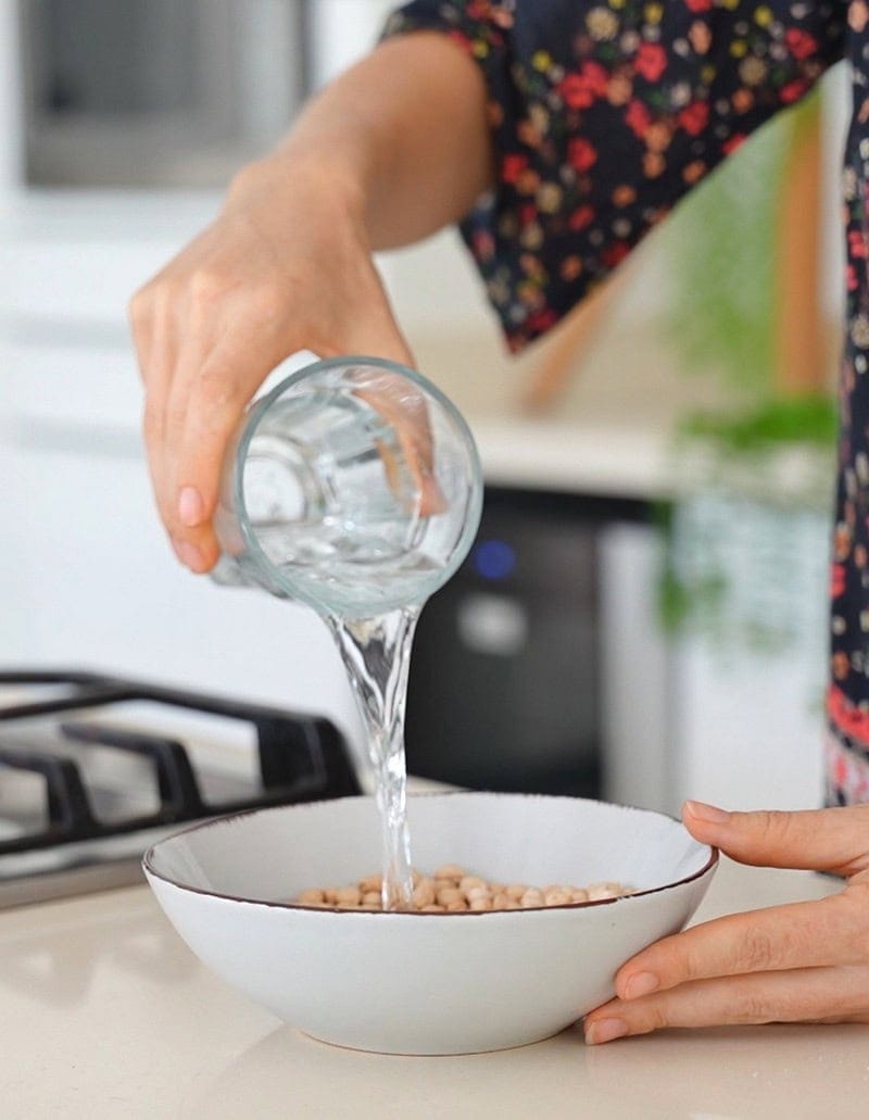 Elavegan pours water from a glass into a bowl of dry chickpeas on a kitchen counter