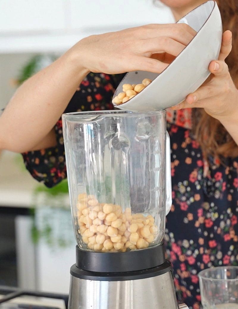 Elavegan pours chickpeas from a white bowl into a blender in a bright kitchen