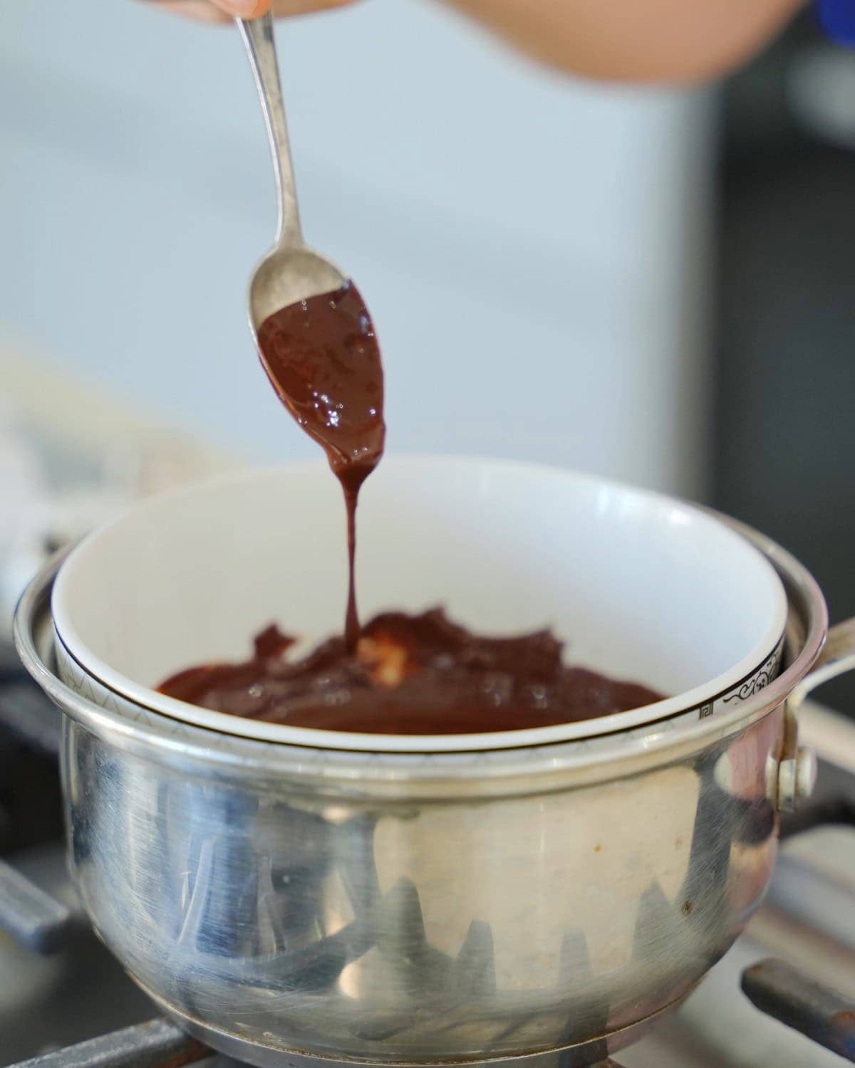Eine Hand hält einen Löffel, von dem geschmolzene Schokolade in eine Schüssel tropft. Die Schüssel steht über einem Topf auf dem Herd, was zeigt, dass die Schokolade geschmolzen wird