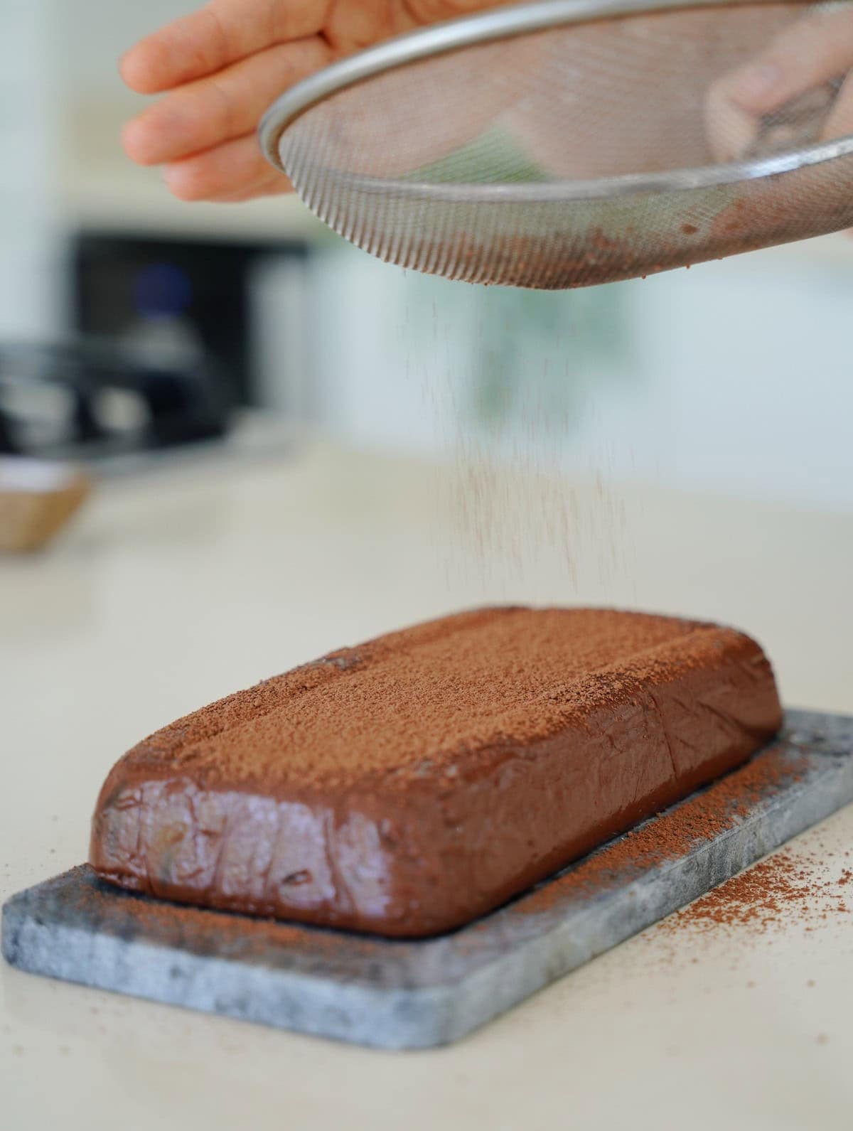 A rectangular chocolate pudding cake on a marble board is dusted with cocoa powder from a sieve