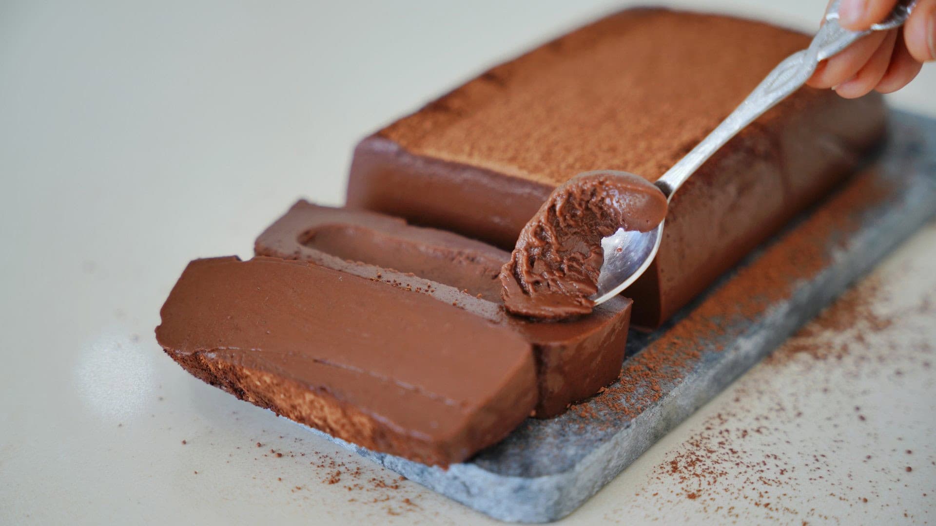 A rich, smooth chocolate pudding loaf is sliced on a stone board. A spoon takes a soft, creamy piece