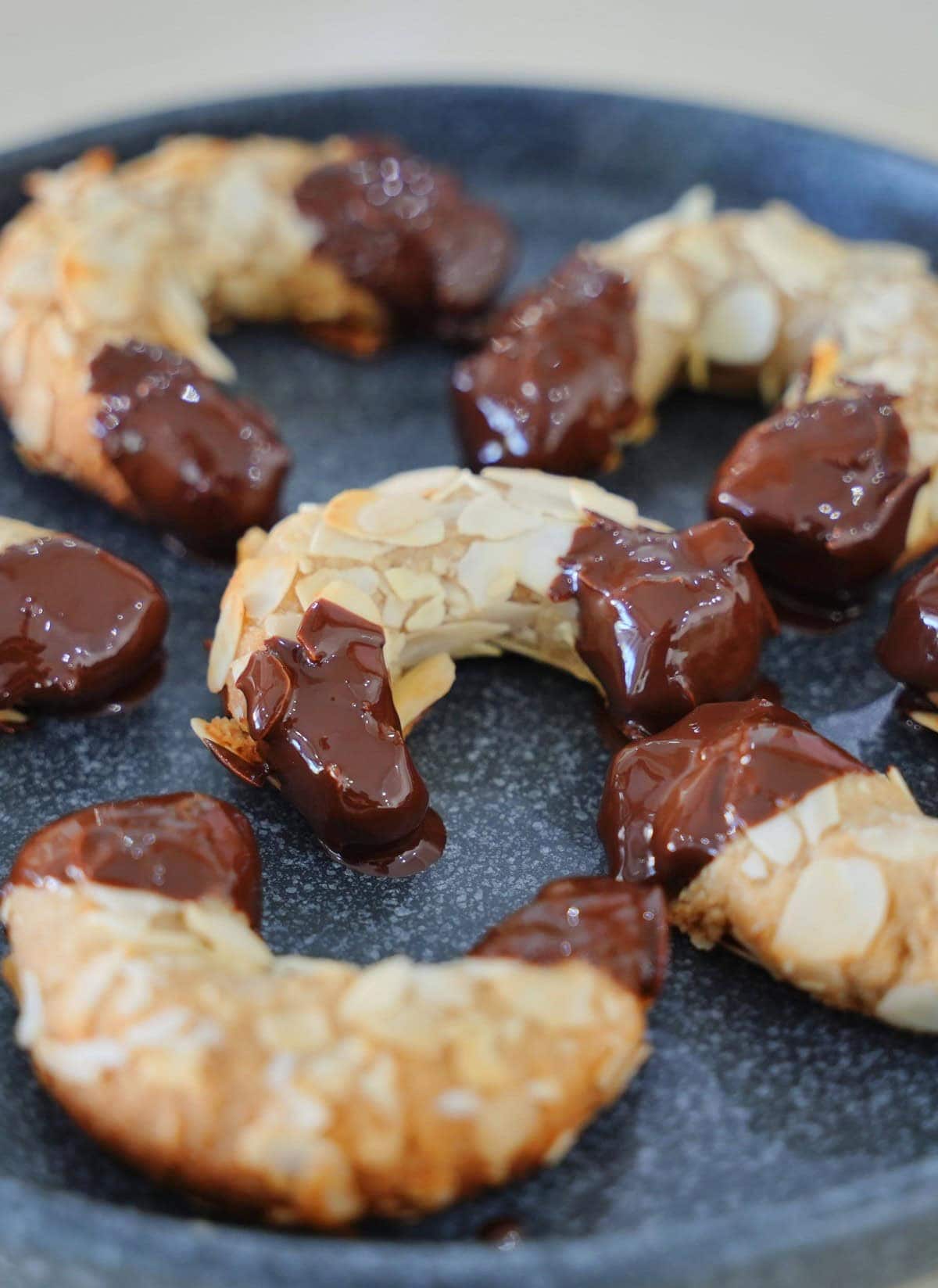 Vegane Mandelhörnchen mit glänzender dunkler Schokolade, angerichtet auf einem dunkelblauen gesprenkelten Teller