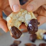 Close-up of hands holding a crescent-shaped cookie coated with almond slices, its ends dipped in melted chocolate