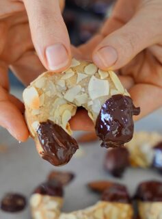 Close-up of hands holding a crescent-shaped cookie coated with almond slices, its ends dipped in melted chocolate