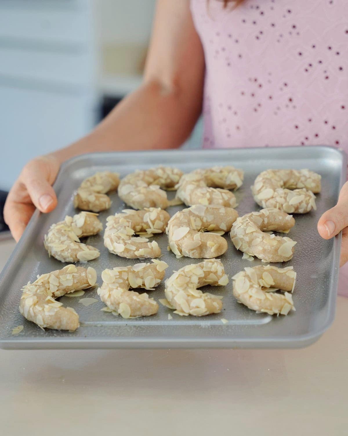Elavegan hält ein Backblech mit halbmondförmigen, mit Mandeln bestreuten Hörnchen