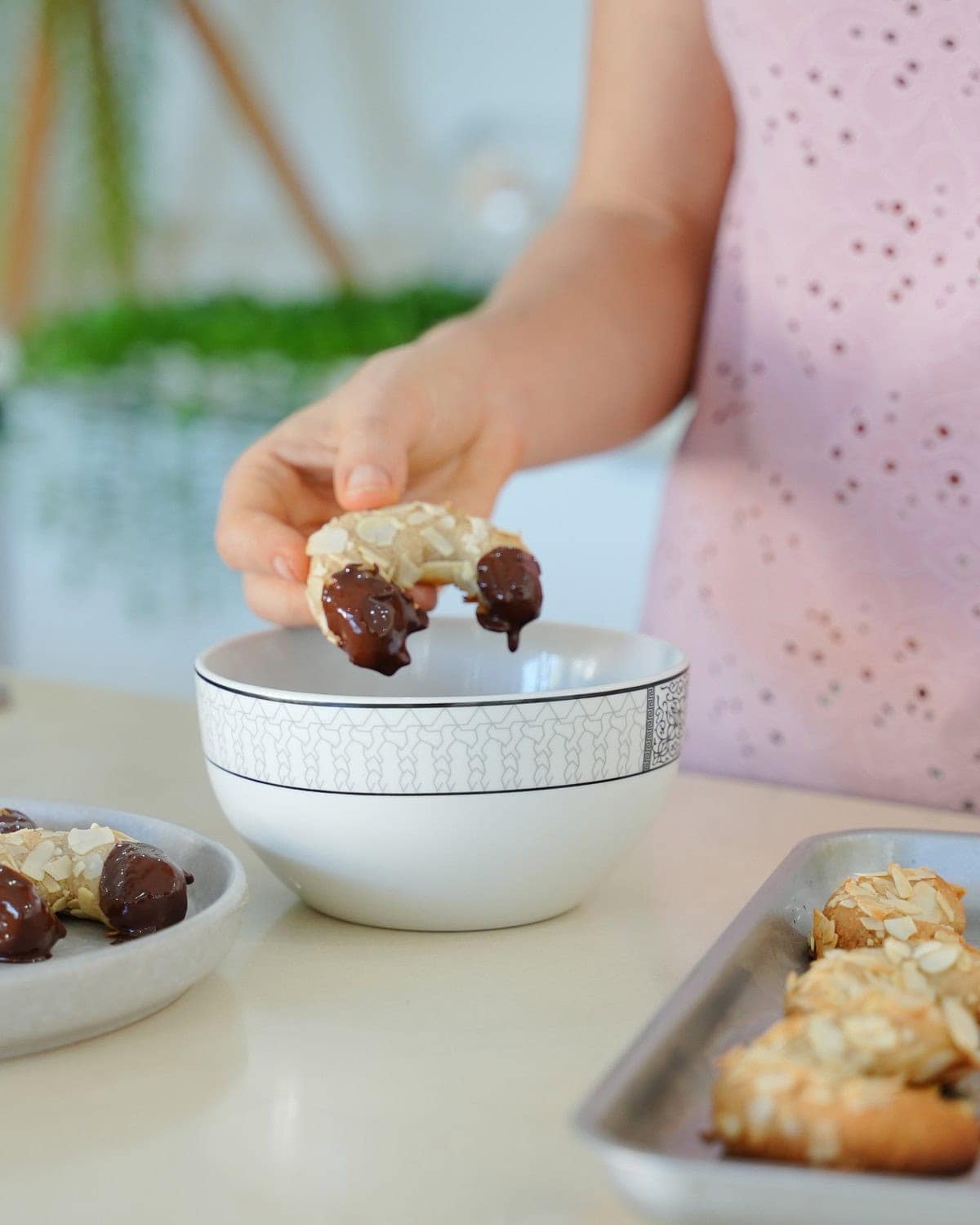 Elavegan hält ein in Schokolade getauchtes Mandelhörnchen über eine dekorative Schale. In der Nähe liegen weitere Mandelhörnchen auf einem Backblech