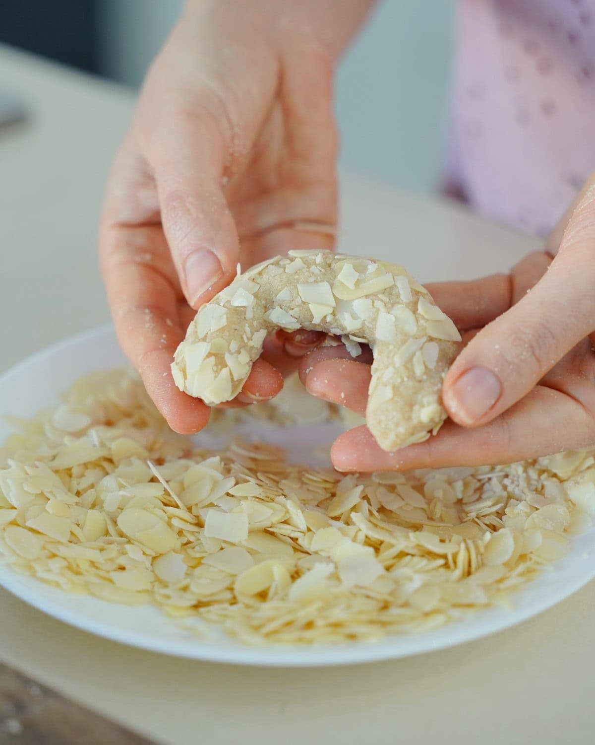 Hände formen ein Mandelhörnchen über einem Teller voller Mandelblättchen