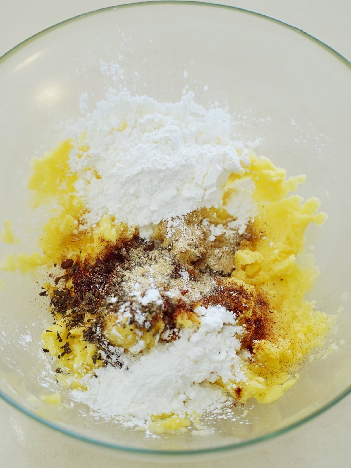 A glass bowl containing mashed yellow potatoes, starch, and sprinkled with various herbs and spices