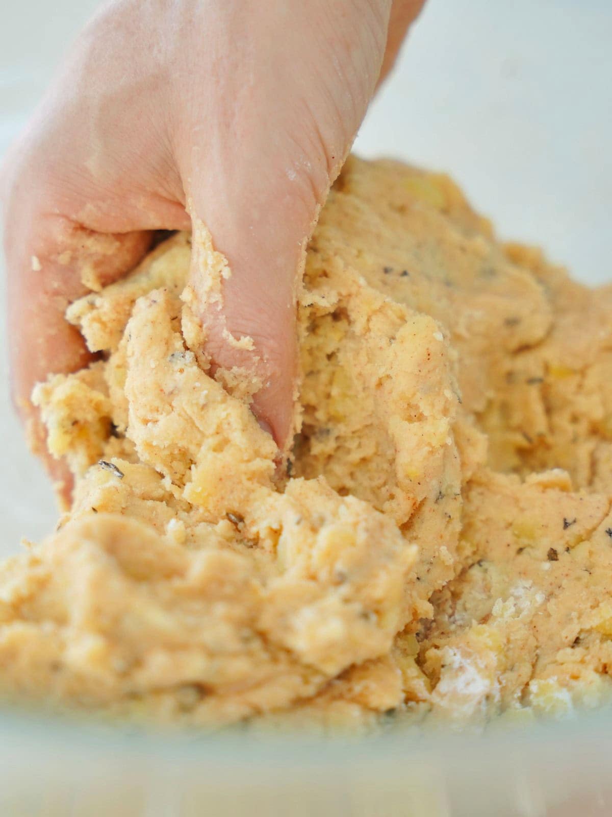 A hand kneads a soft, yellowish dough with visible herbs in a mixing bowl