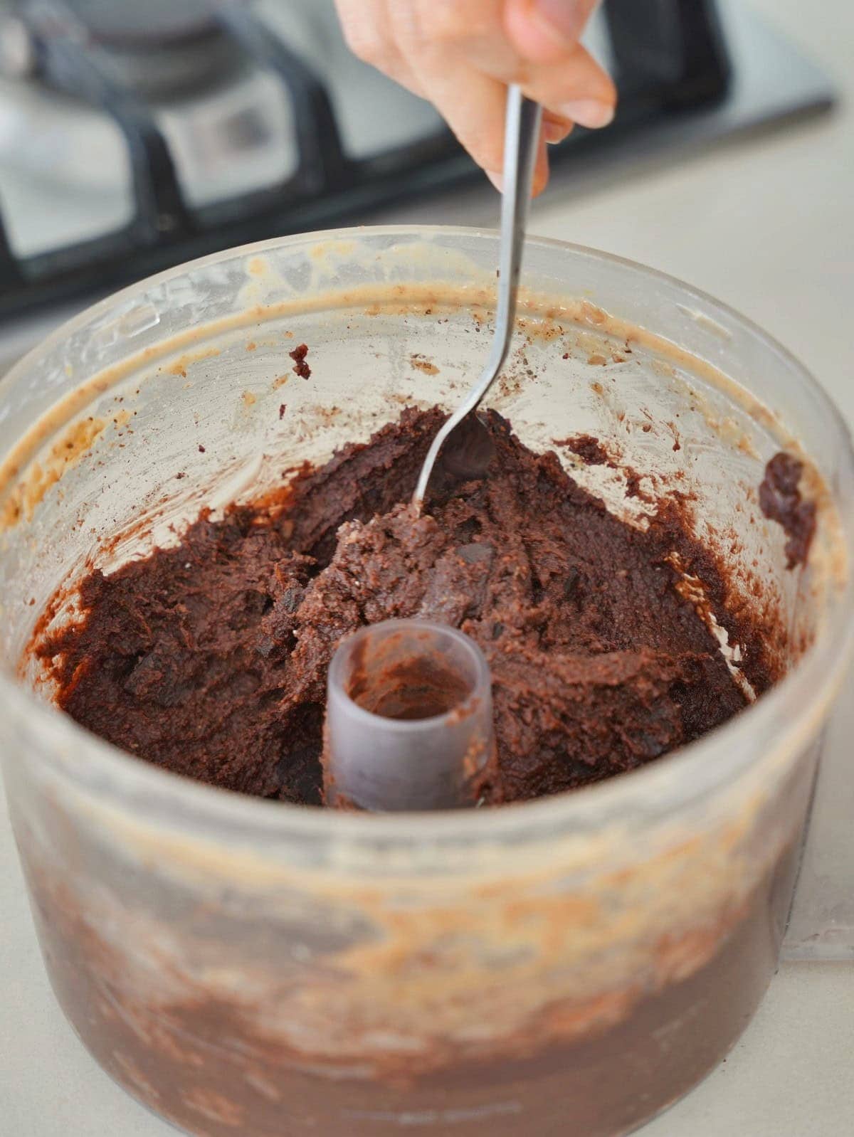 A hand stirs thick chocolate brownie batter in a clear food processor with a spoon