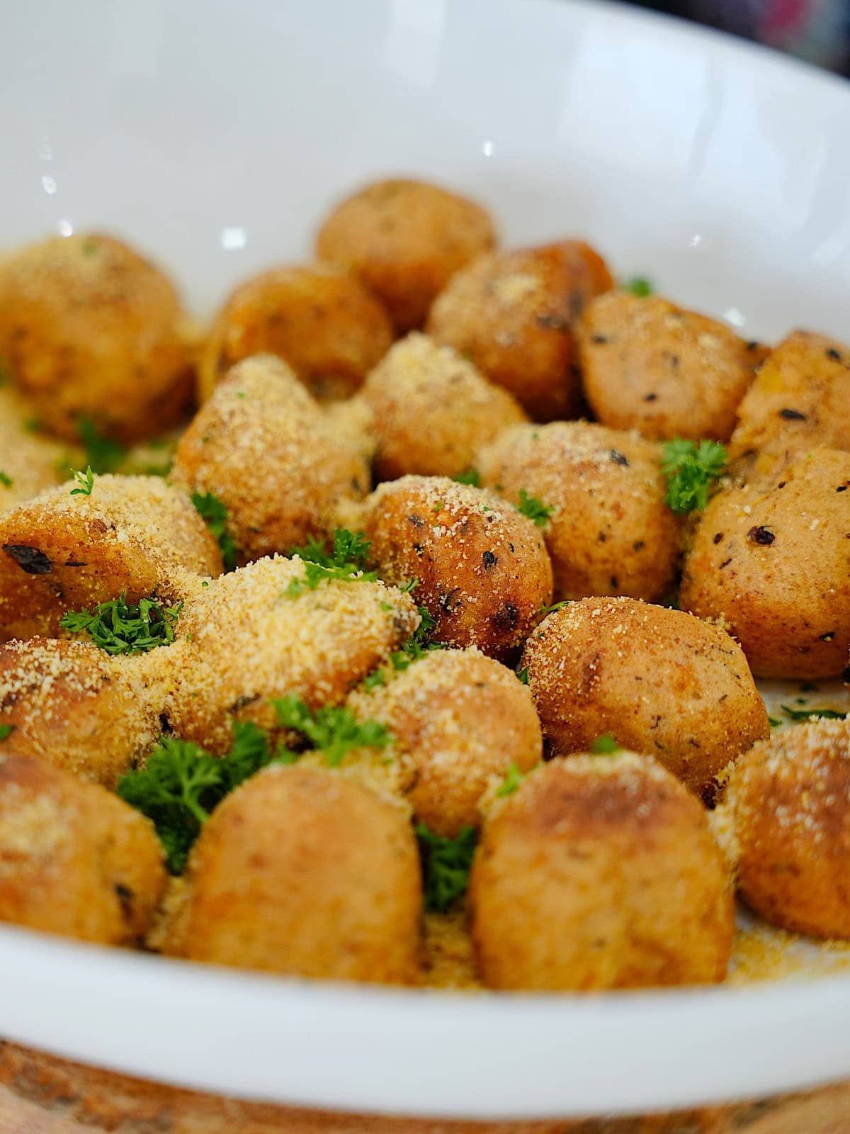 A white bowl filled with golden-brown, crispy potato balls, sprinkled with vegan Parmesan and parsley