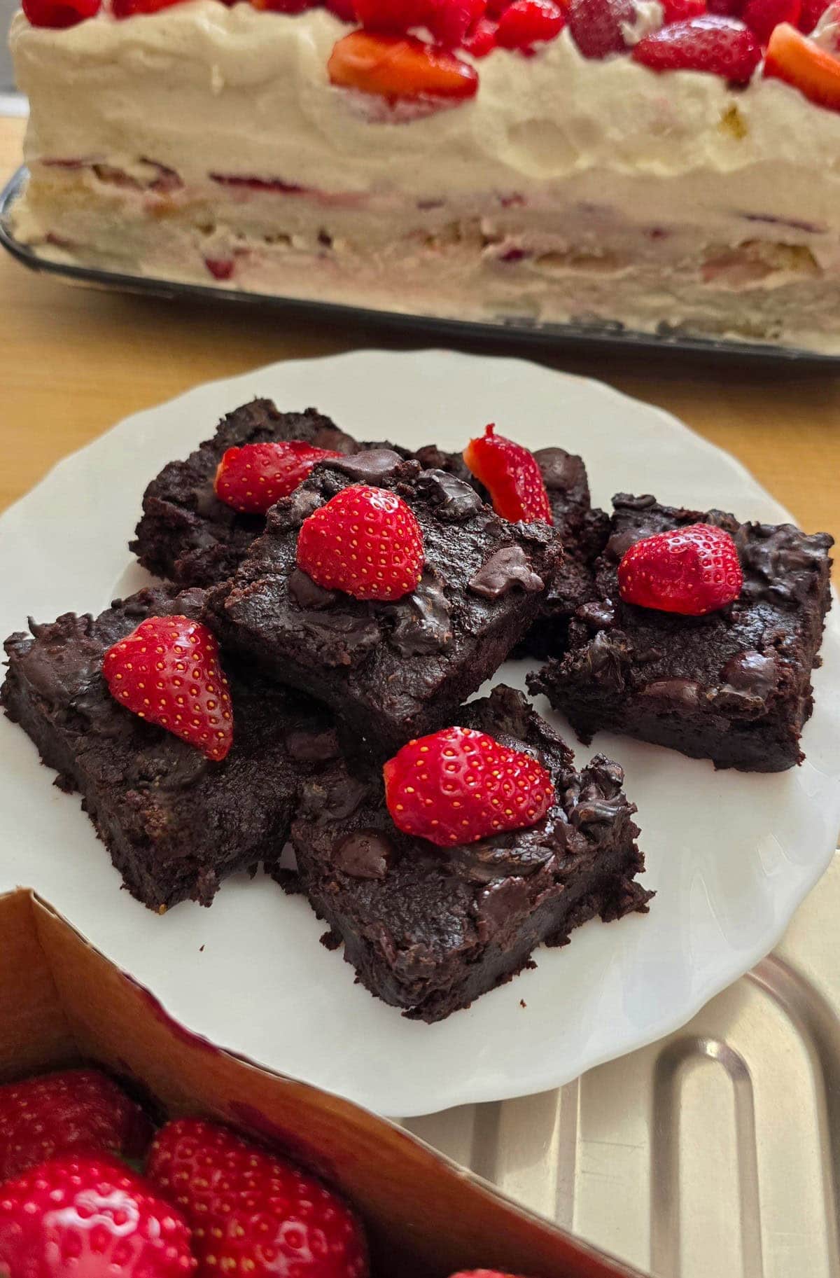 Chocolate brownies topped with fresh strawberries are displayed on a white plate. In the background, there's a creamy cake