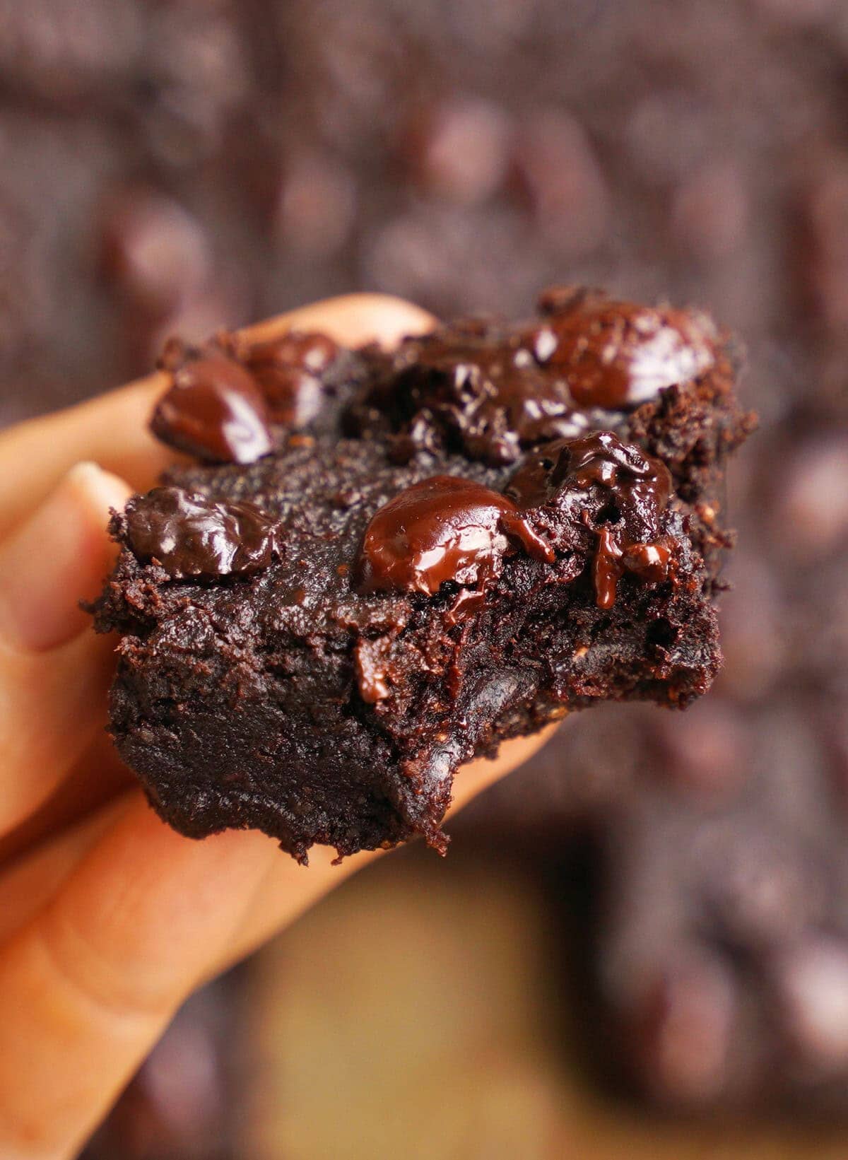 Close-up of a hand holding a gooey chocolate date brownie with melted chocolate chips on top