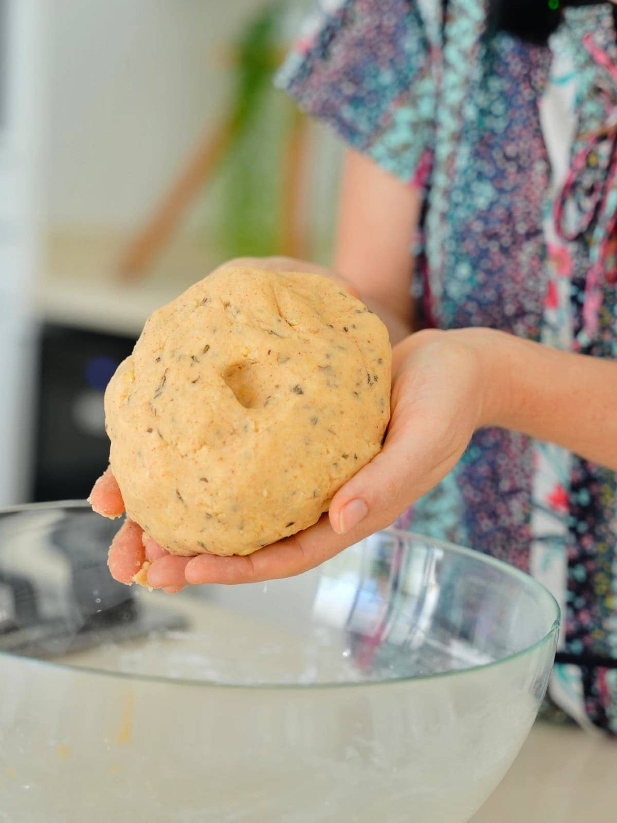 Elavegan holds a ball of dough with herbs, over a glass bowl in a kitchen