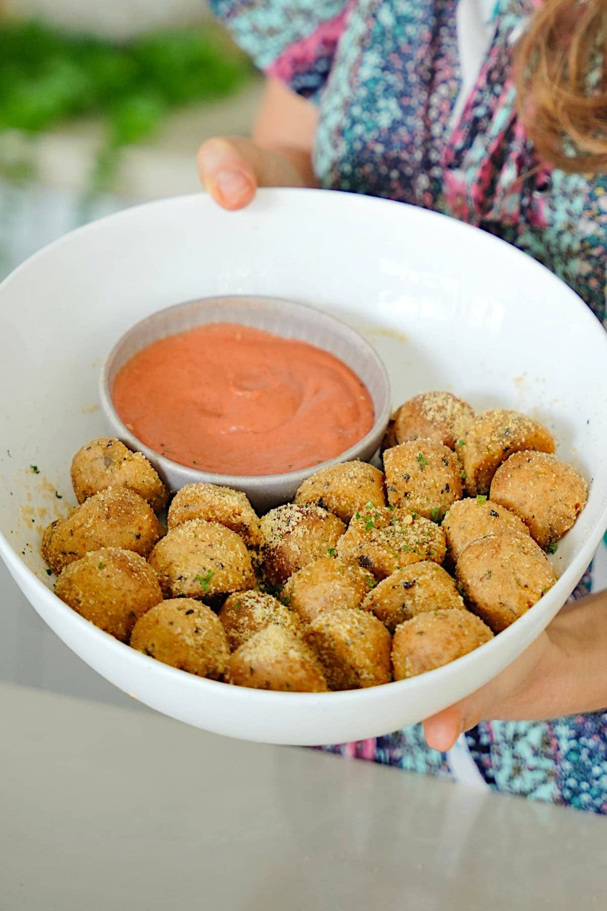 Elavegan holds a white bowl filled with air fryer potato balls topped with herbs next to a dip
