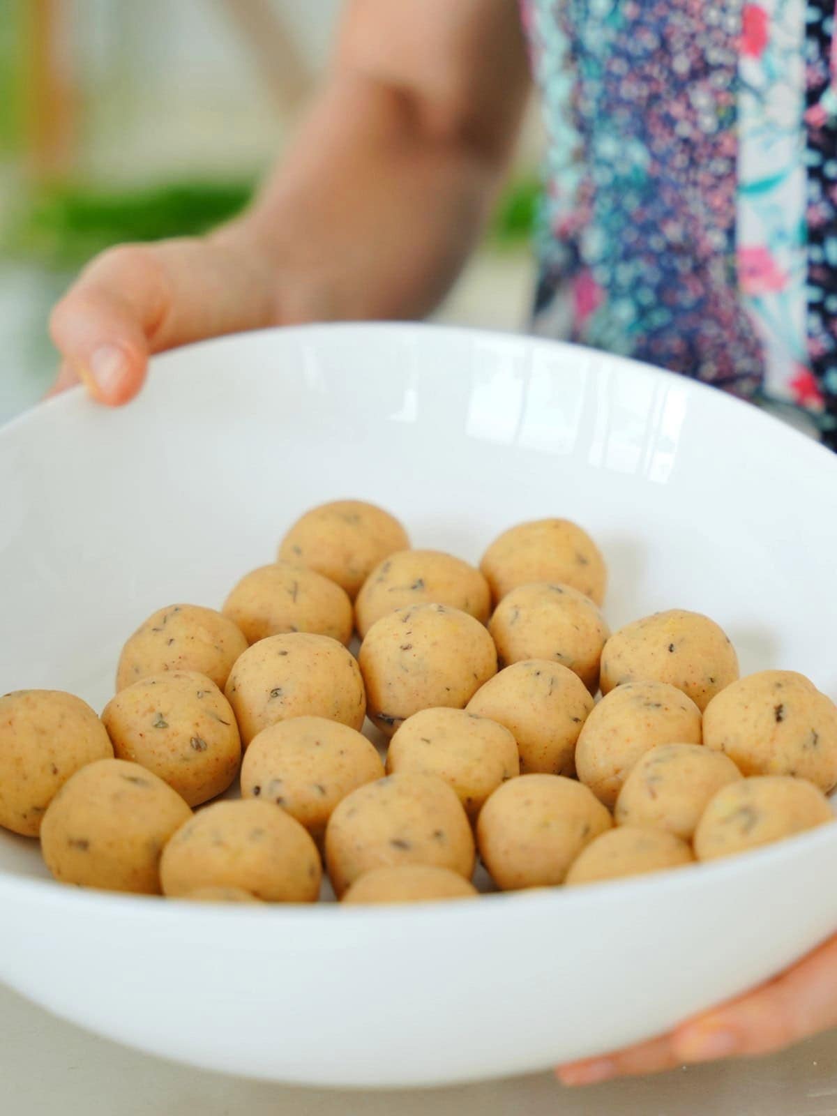 Elavegan holds a white bowl filled with round, beige dough balls speckled with herbs