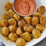 Golden-brown seasoned crispy potato balls in a white bowl, surround a ramekin of creamy, orange dipping sauce