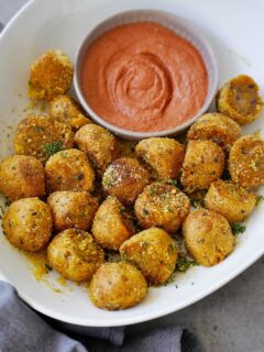 Golden-brown seasoned crispy potato balls in a white bowl, surround a ramekin of creamy, orange dipping sauce