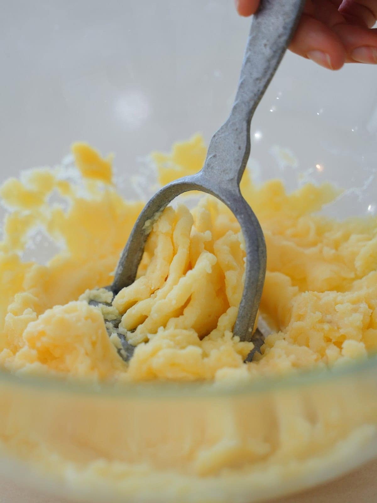 Hand holding a masher, pressing creamy mashed potatoes in a clear glass bowl
