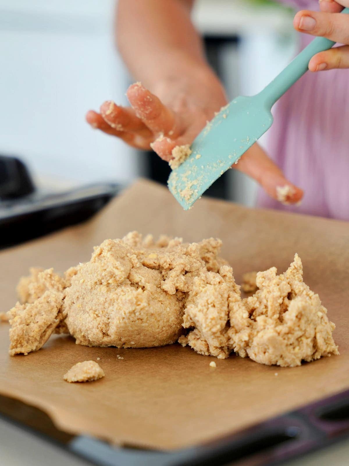 Hands using a teal spatula to spread crumbly chickpedough on parchment paper